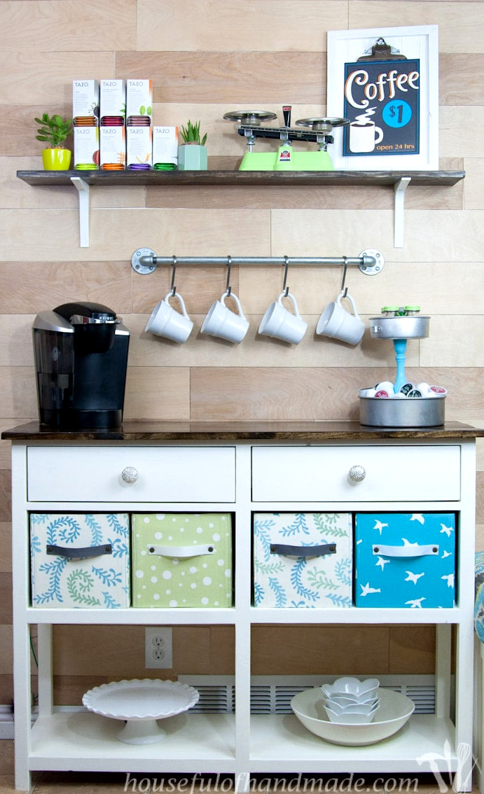 Coffee bar created on a white coffee station table with a shelf above it, hanging rack for mugs, fabric storage boxes, and k-cup storage. 