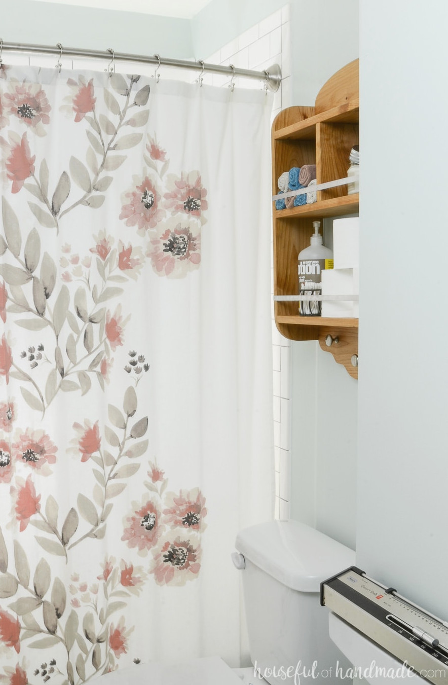 Wood shelves above the toilet holding toilet paper and other bathroom essentials. Positioned beside a watercolor floral shower curtain. 