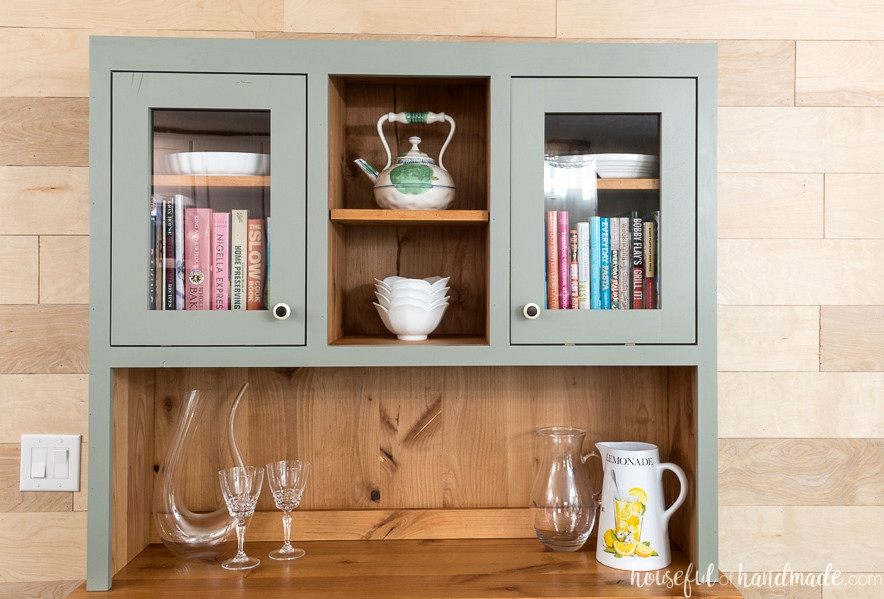 Natural wood hutch with a painted gray interior holding platters, cookbooks, and baking dishes.