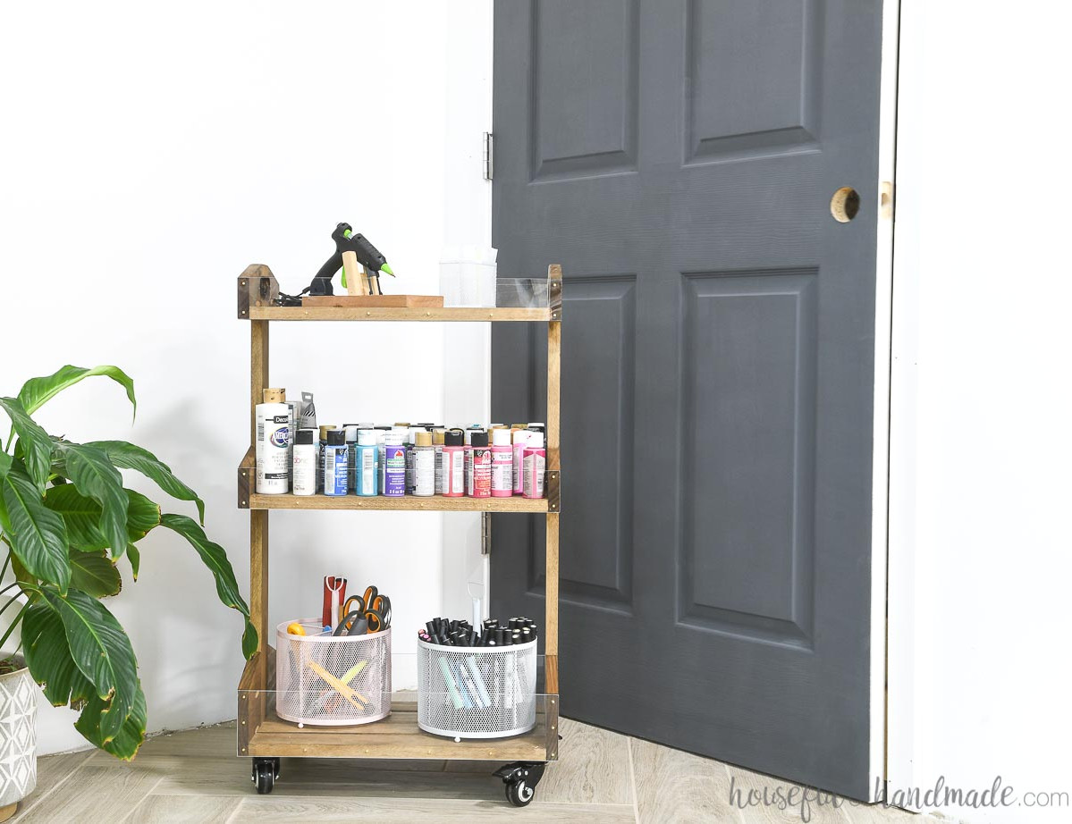 Wooden rolling cart filled with craft supplies positioned in front of a charcoal-painted door.