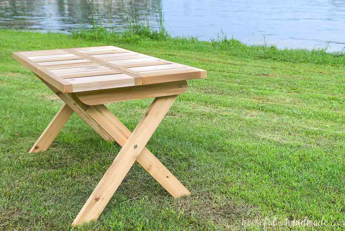 X-leg folding picnic table built from wood and positioned on the lawn beside a lake. 