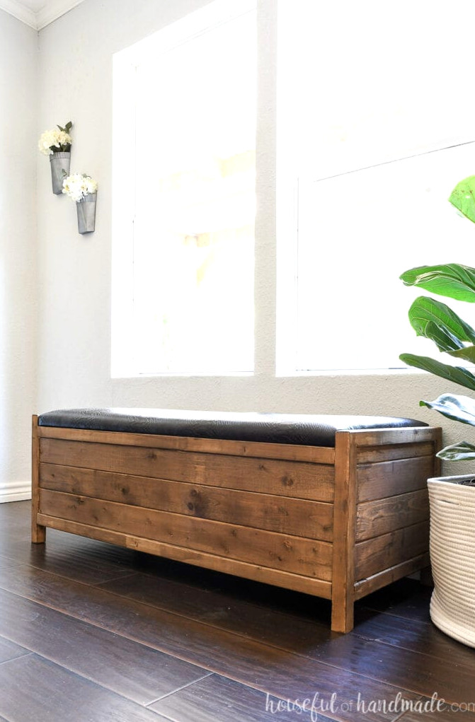 Upholstered storage bench featuring a black faux-leather top, placed near a window beside a fiddle leaf fig plant.