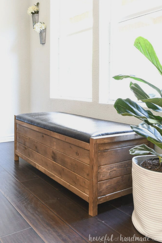 Brown and black farmhouse-style storage bench positioned against pale gray walls beside a fiddle leaf fig tree.