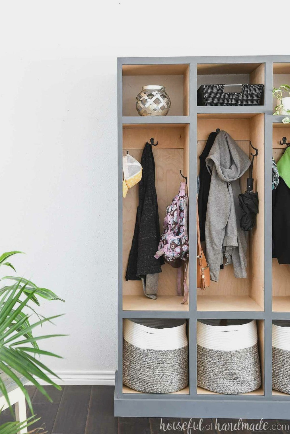 View of one half of the mudroom storage lockers with sweaters and backpacks suspended from the hooks and a fern in the foreground.
