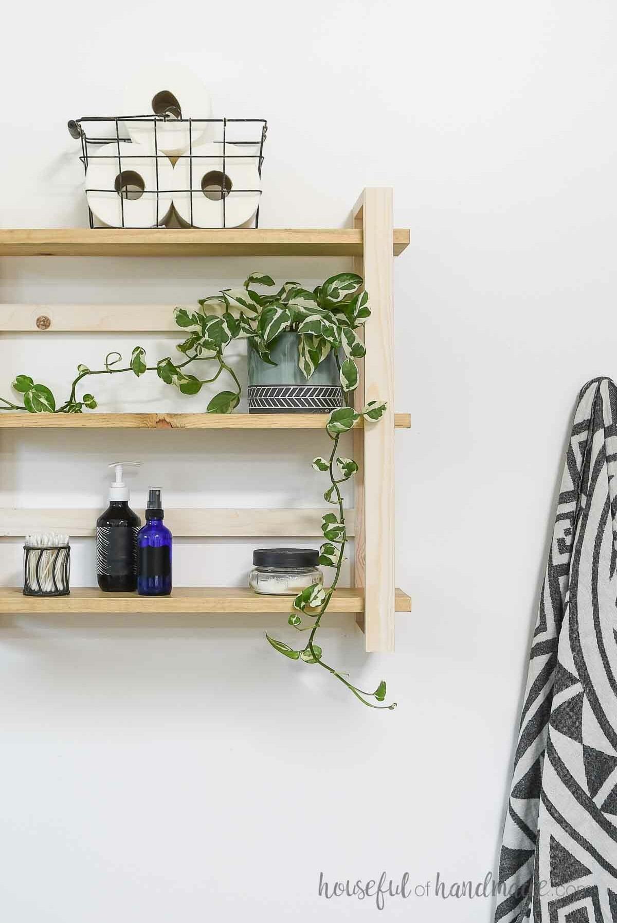 Three-tier wall shelves mounted on a white wall beside a towel hanging from a hook.
