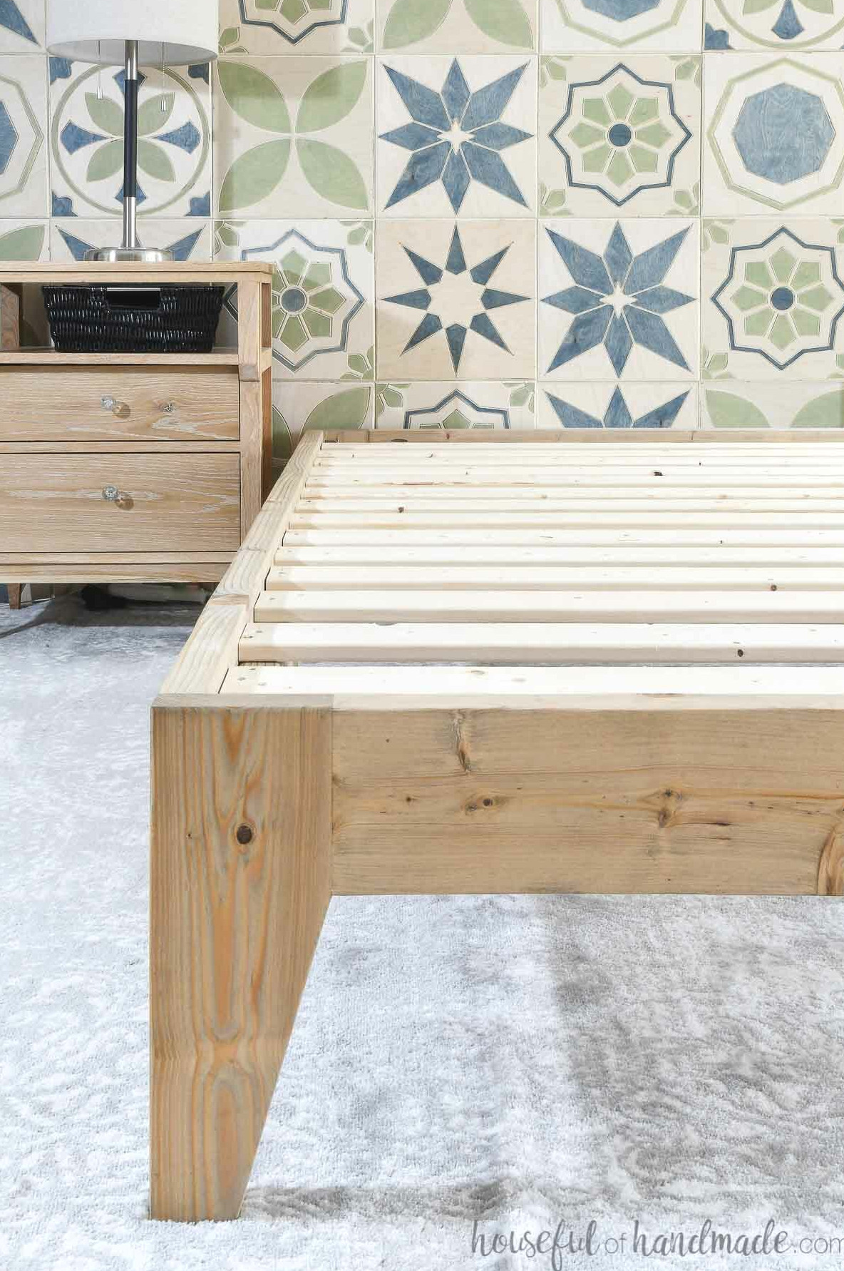 DIY platform bed frame with no mattress on top, placed in a bedroom featuring a stained tile accent wall.