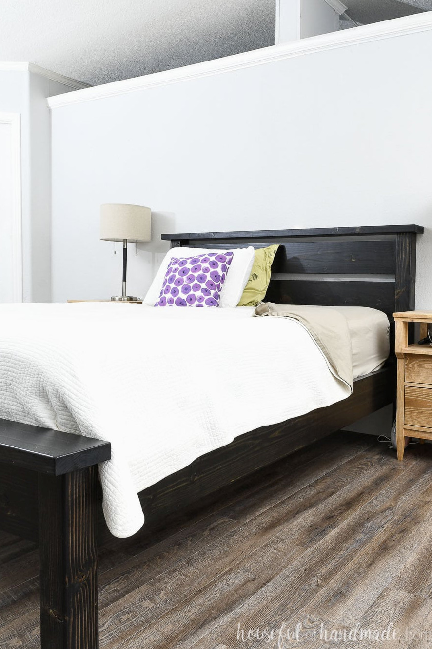 DIY Queen bed frame built from construction lumber, stained black, and styled with white bedding and bright throw pillows.