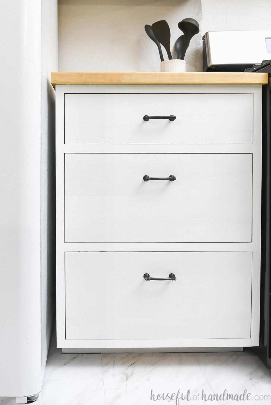 White drawer base cabinet with 3 drawers, one smaller on top and two larger on bottom, with a wood countertop.