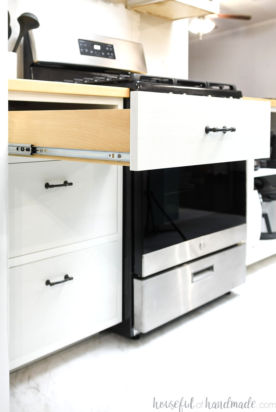 White drawer base cabinet next to a range that has the top drawer open showing the drawer slides on the side.