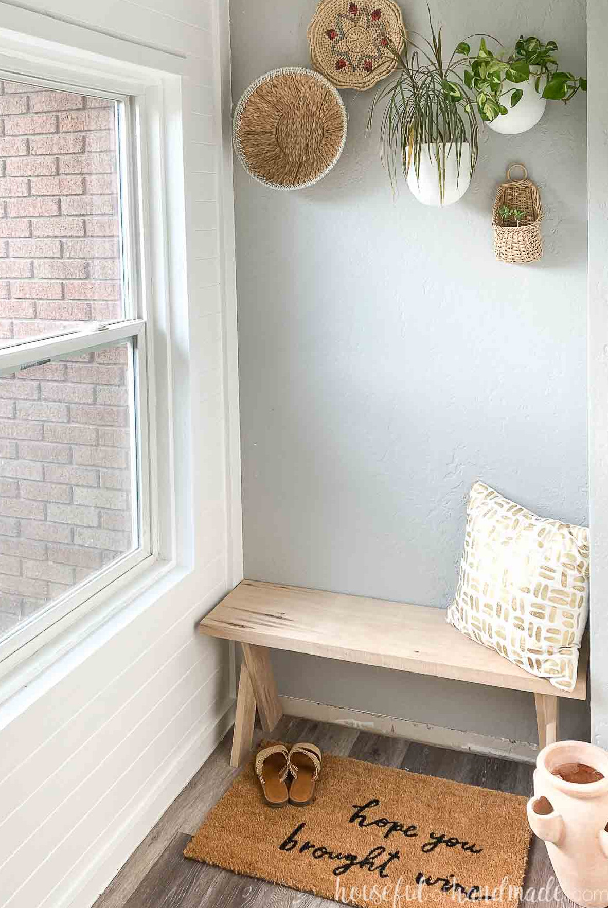 Entryway featuring an x-leg bench made from light wood, a gold and white pillow, adobe planter, rug, and sandals nearby. 