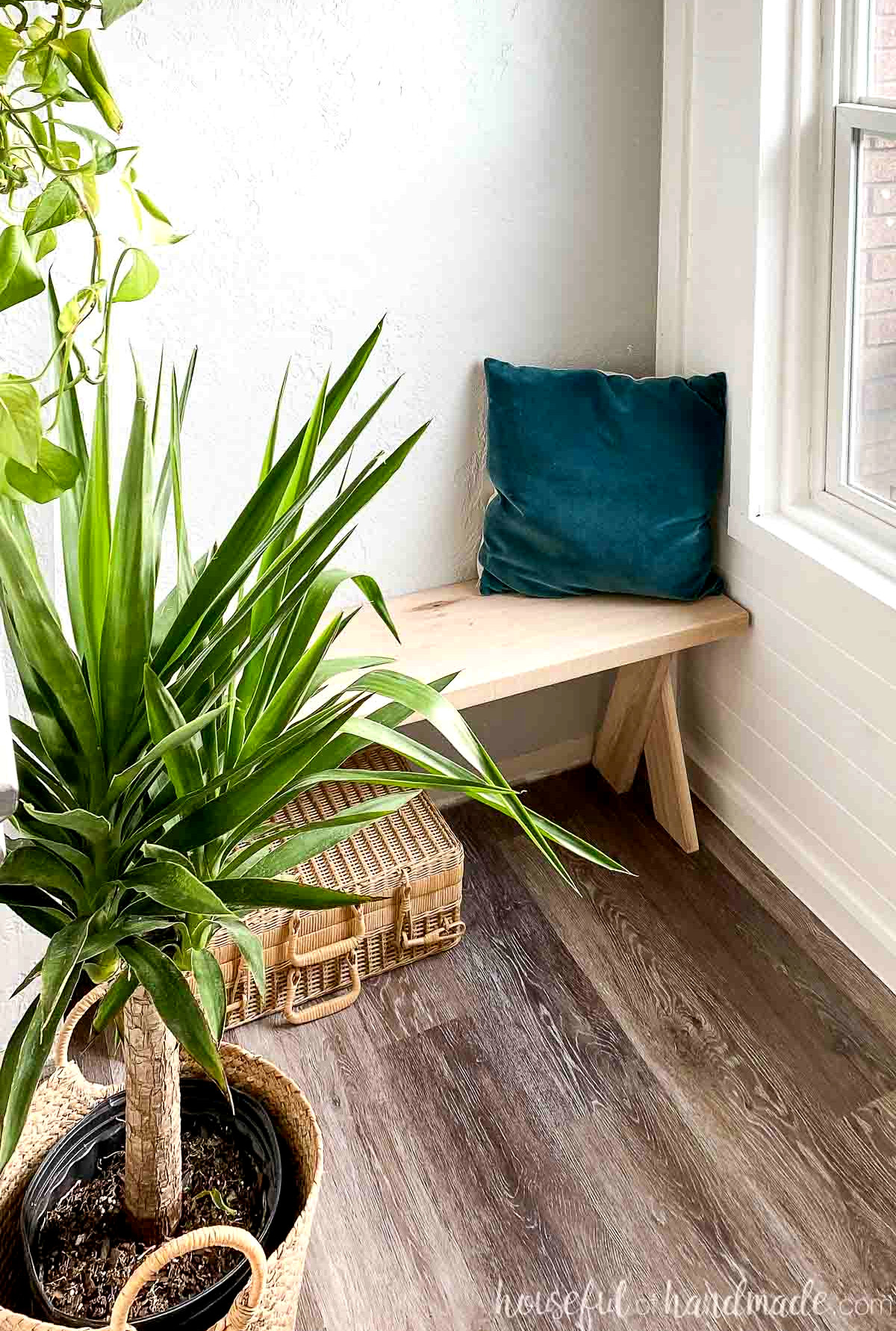 Tropical potted plant in front with the DIY x-leg bench behind it, topped with a velvet pillow. 