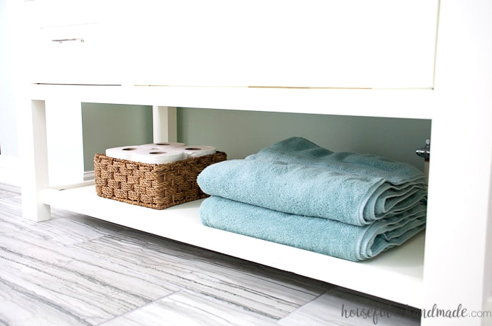 Wide open shelf at the base of a DIY vanity holding towels and toilet paper. 