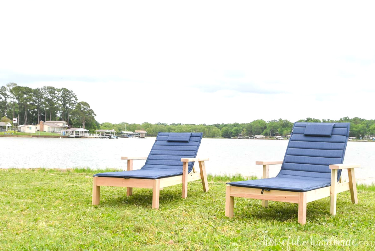 Two pool lounge chairs with thin cushions on the lawn by a lake.