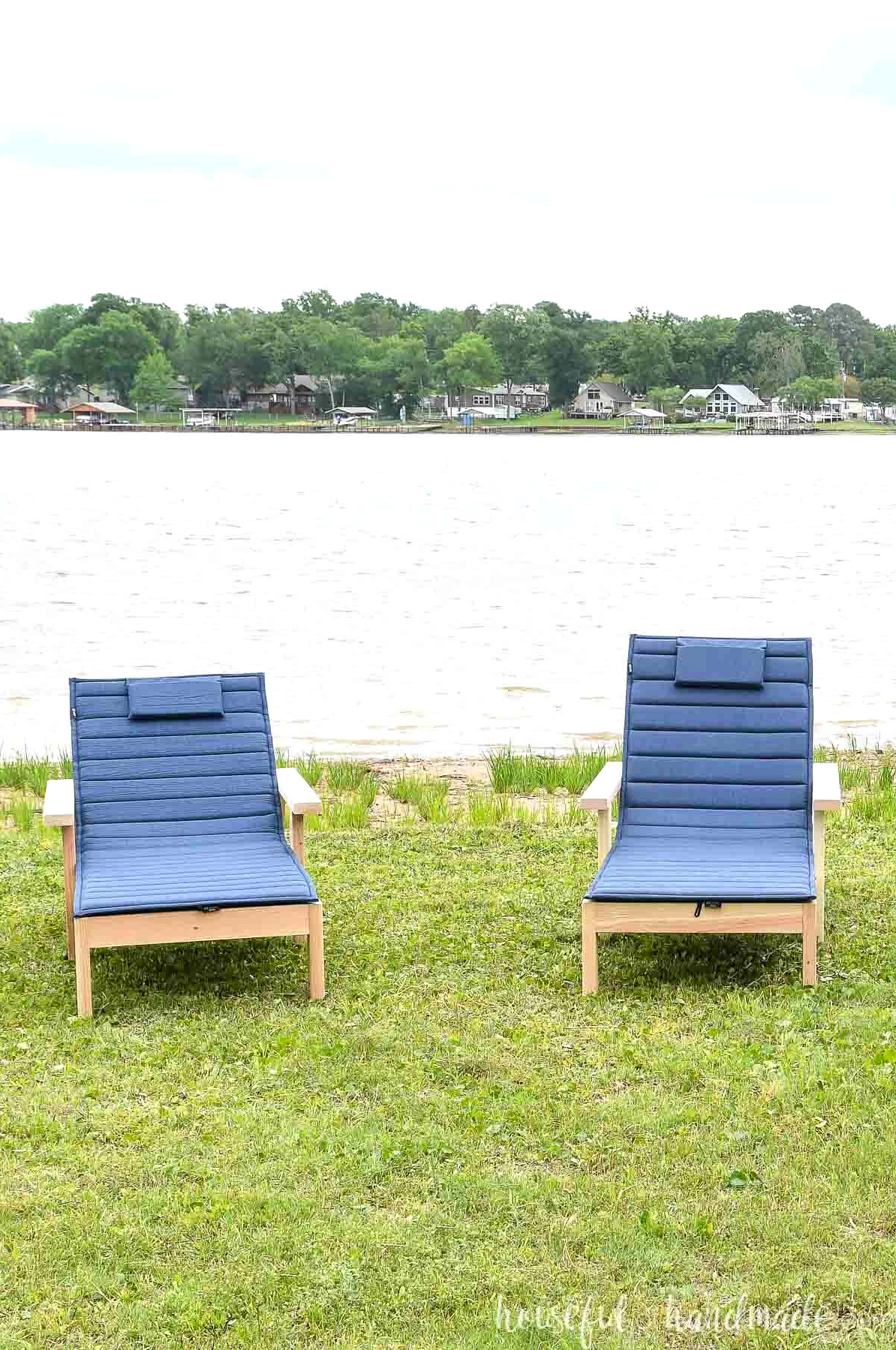 Two lounge chairs with thin, roll-up cushions, sitting in the grass by a lake.