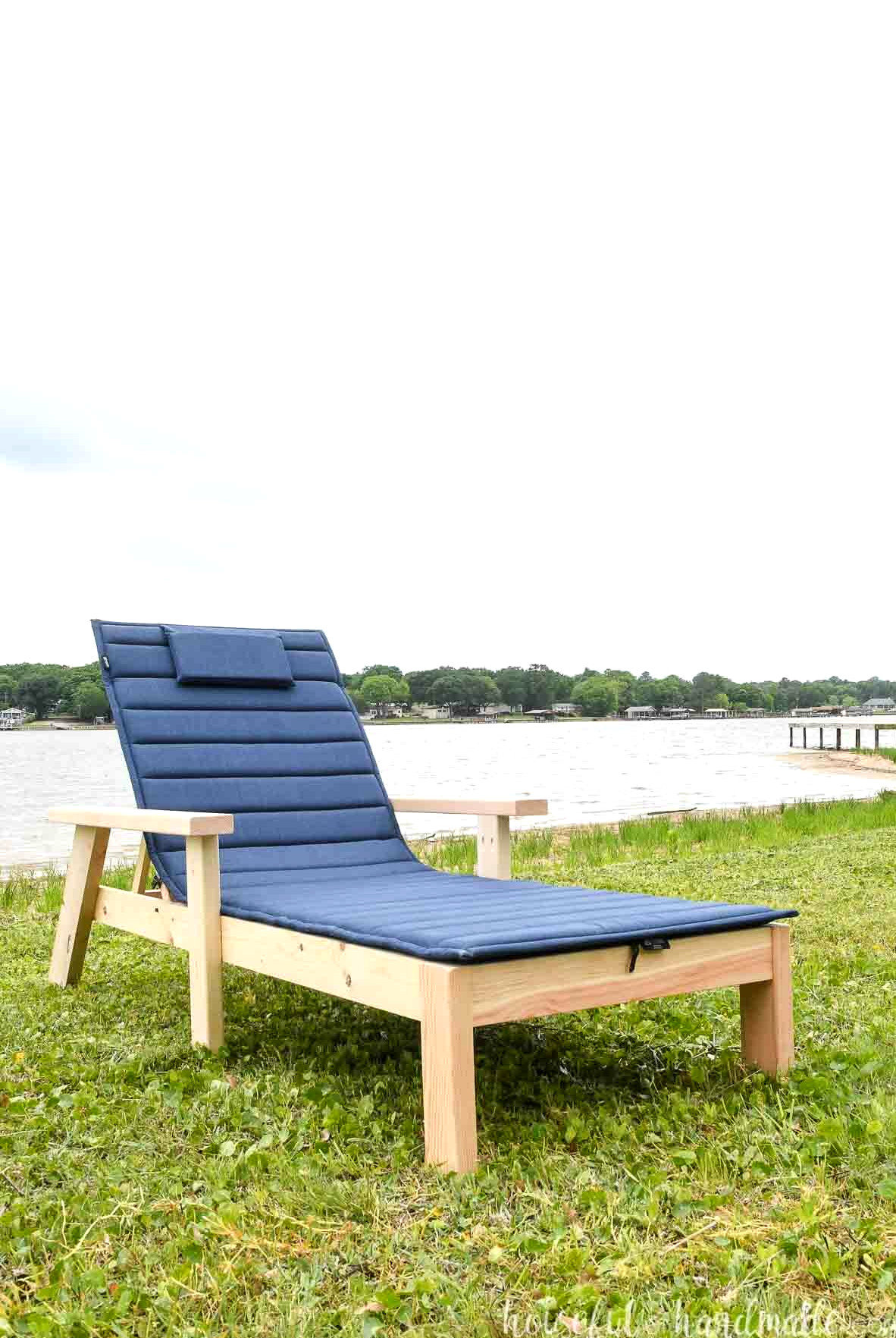 Wood chaise lounge chair with navy cushion on a lawn in front of a lake.