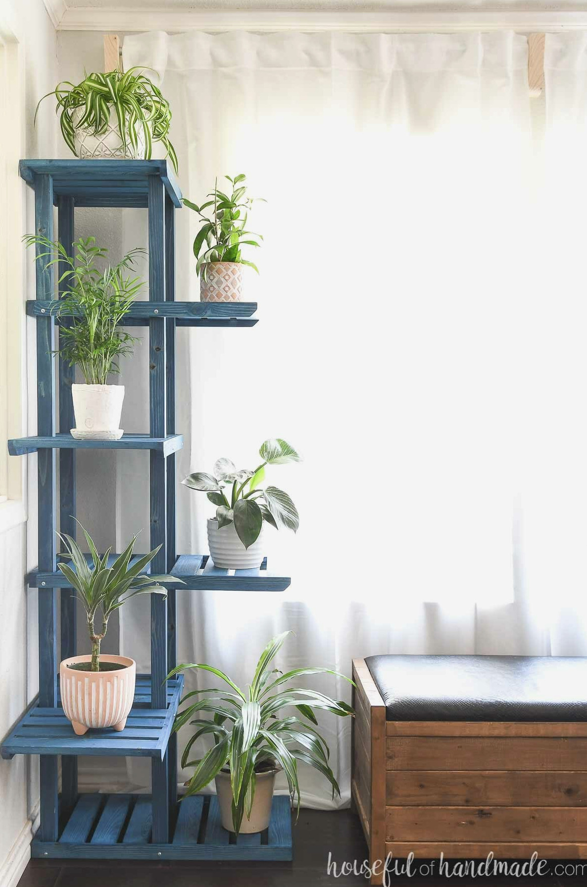 Tall corner plant stand filled with indoor houseplants, positioned beside a bench in front of a window. 