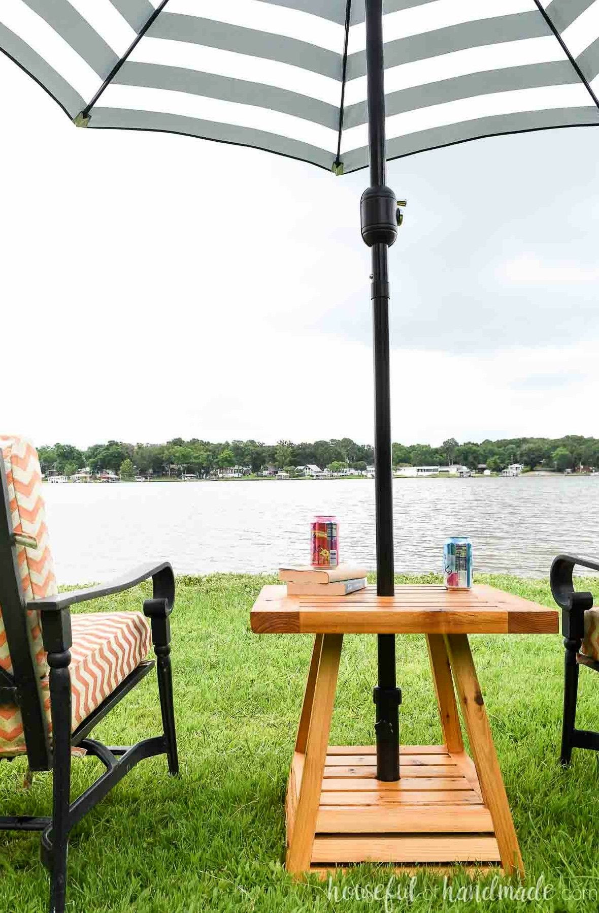Umbrella side table with a black and white striped umbrella in it next to black frame outdoor chairs in front of a lake.