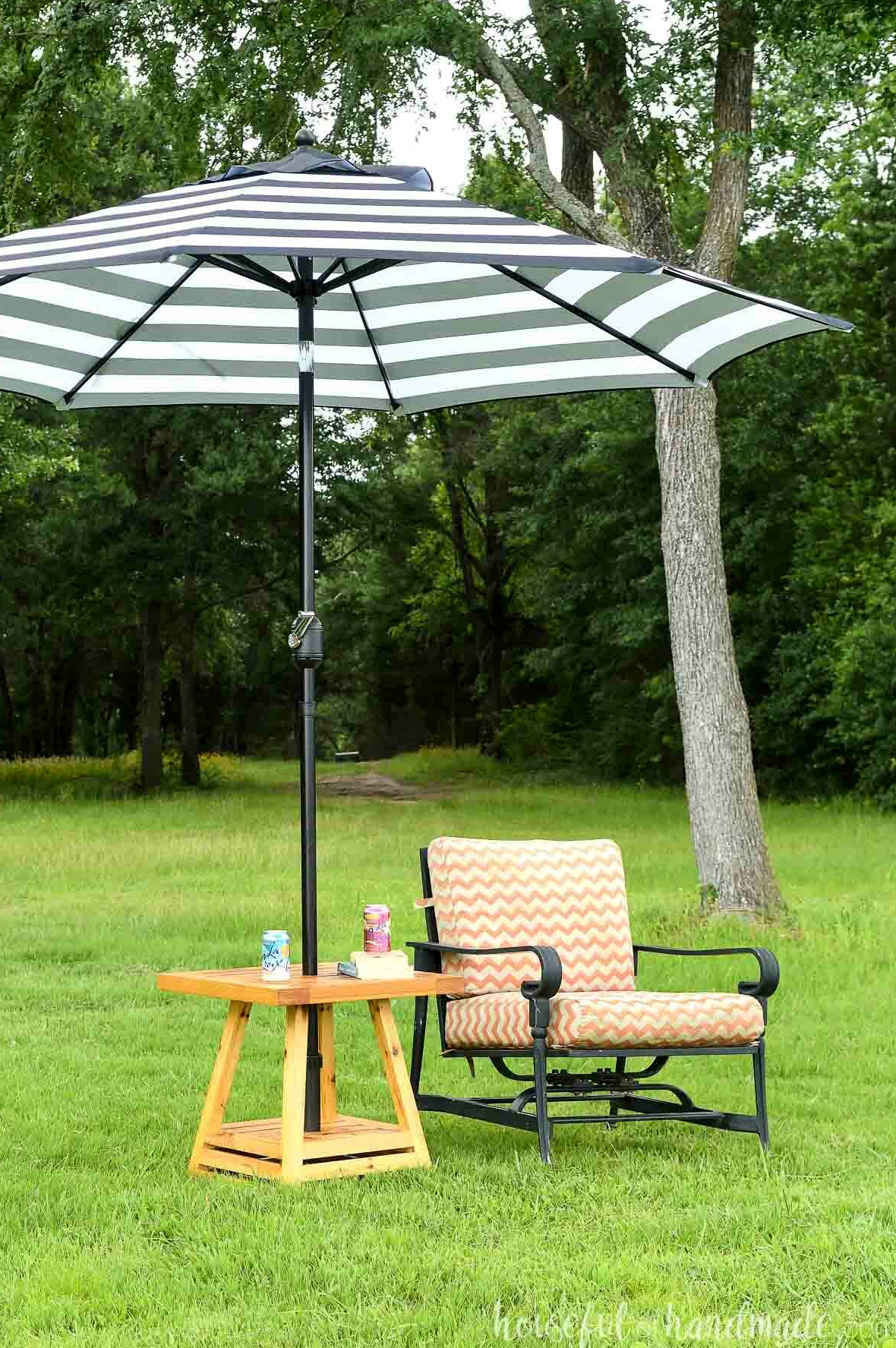 Small outdoor umbrella table next to chair on a lawn with trees in the background.