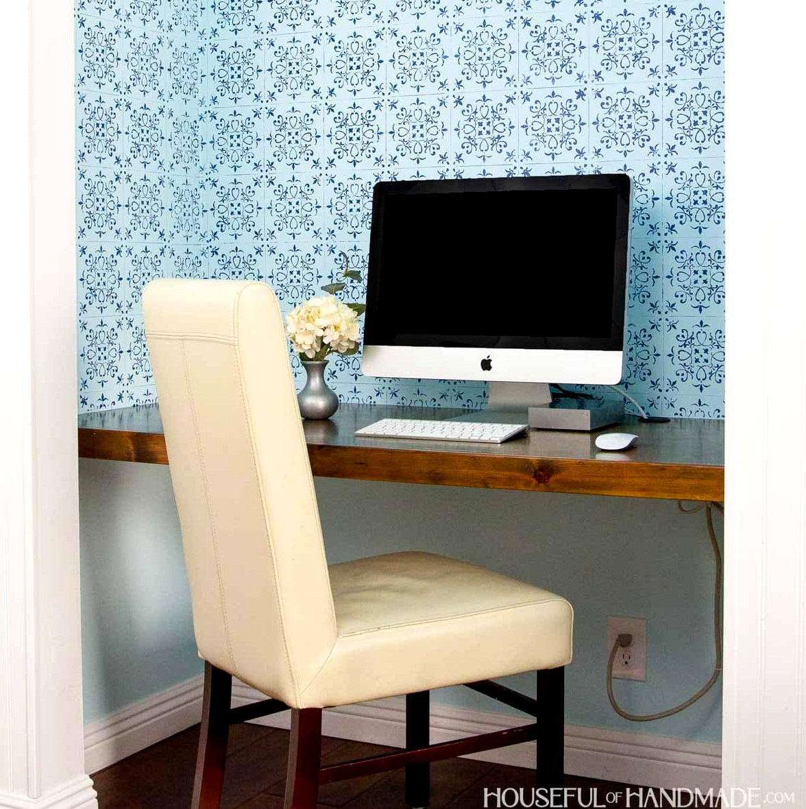Office in closet shown with wood desk, office chair, blue wall paper and iMac computer.