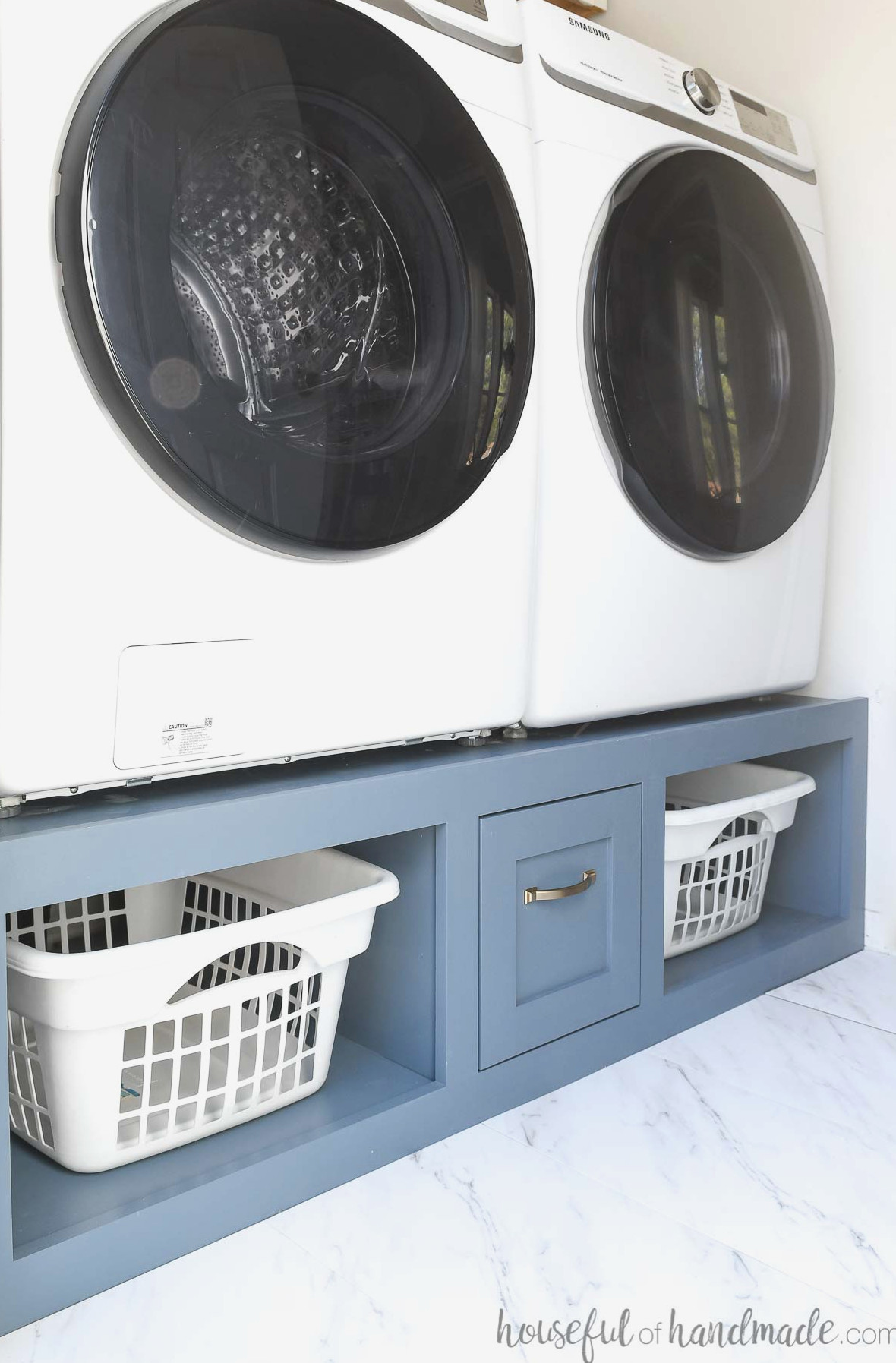 Laundry room featuring a washer and dryer placed on a blue-painted base with laundry basket shelves and a drawer.