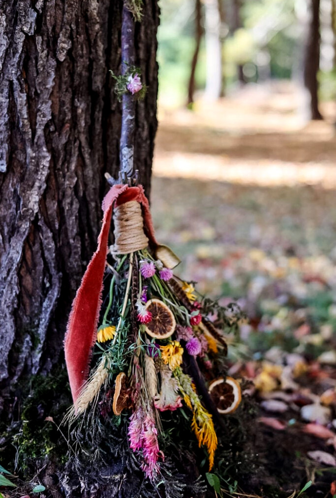 a witch broom made with flowers, greenery, herbs, and orange slices leaning against a tree