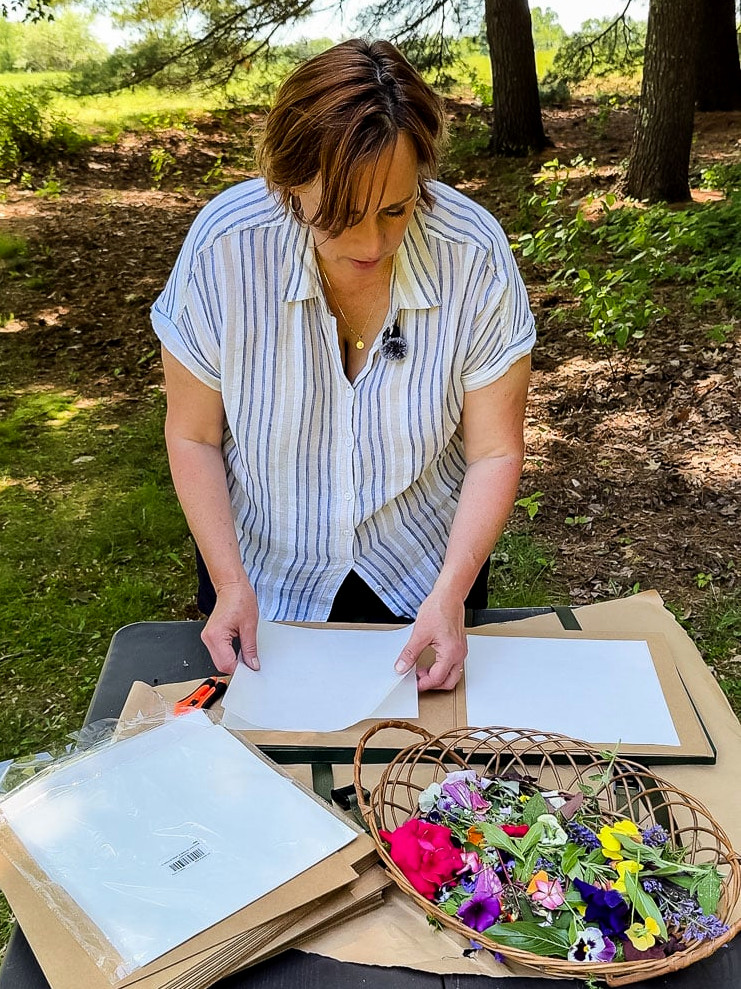 woman using a flower press outside with trees behind her and a field