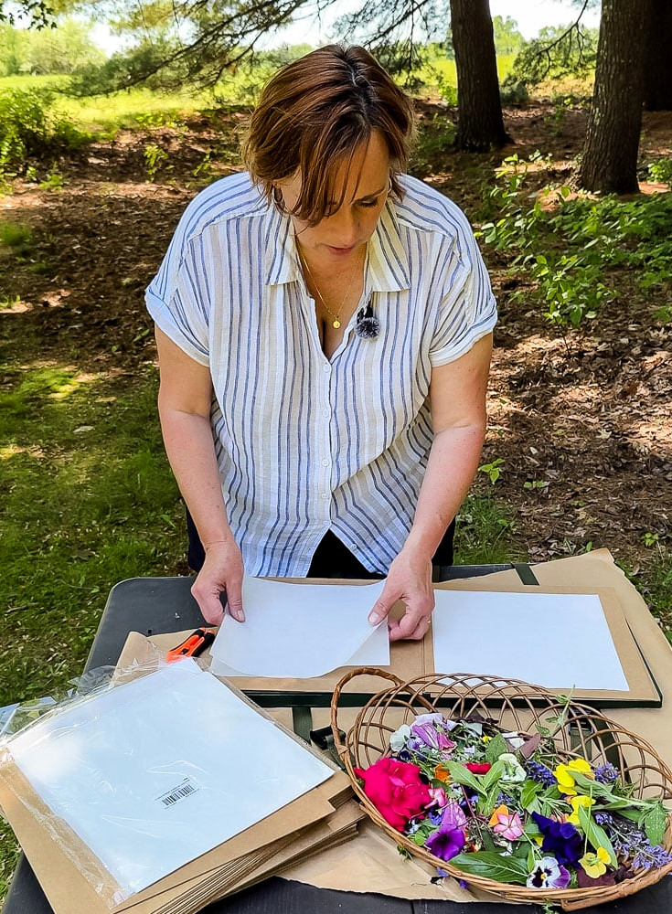 woman using a flower press outside with trees behind her and a field